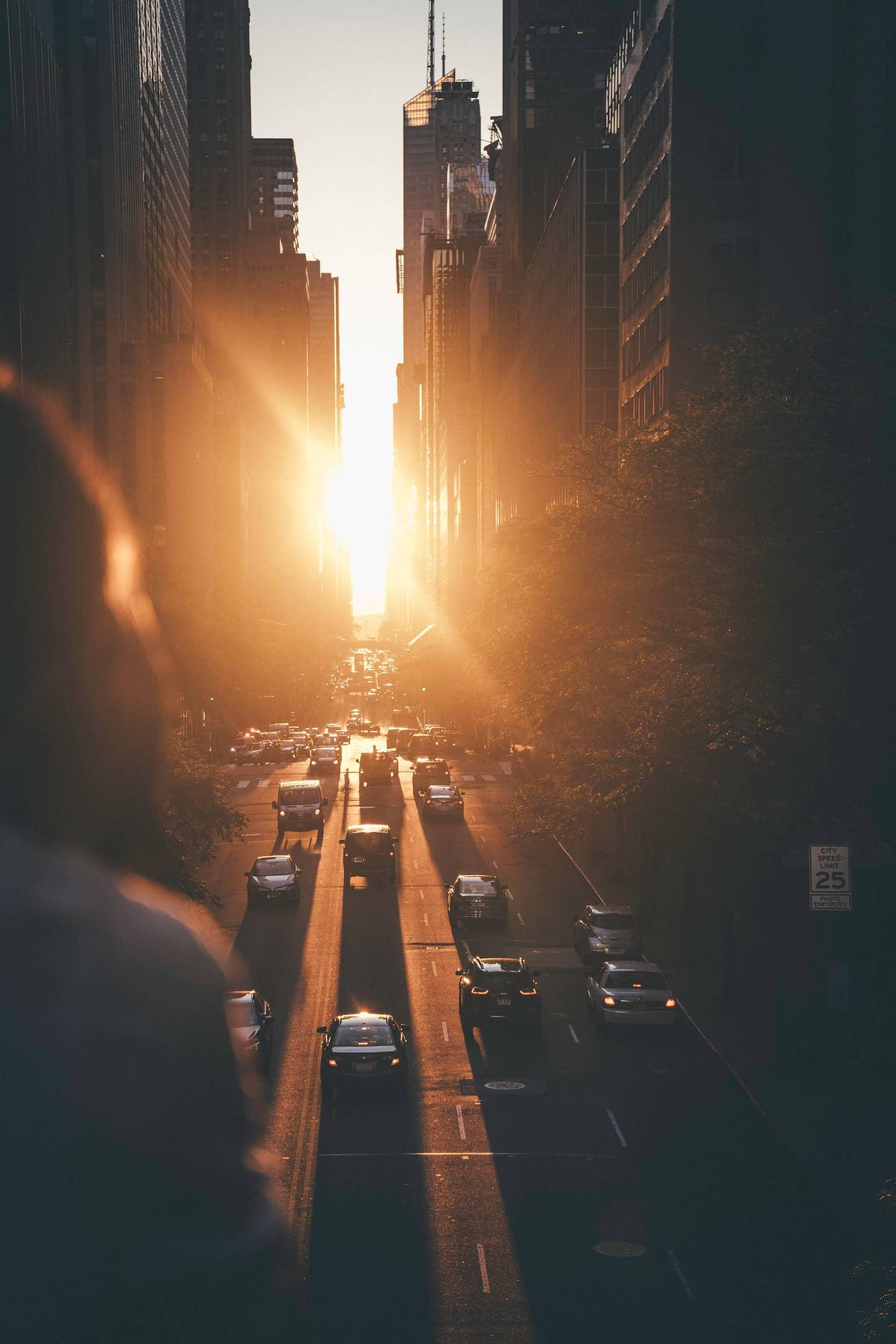 A person standing on a bridge looking towards the sunset as the rays peek through skyscrapers upon a busy road in New York City